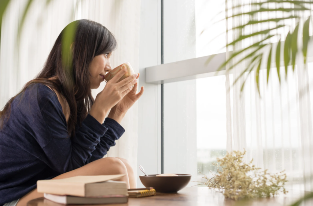 Woman sitting by a window with a warm drink, taking time to relax and prioritize her mental health.